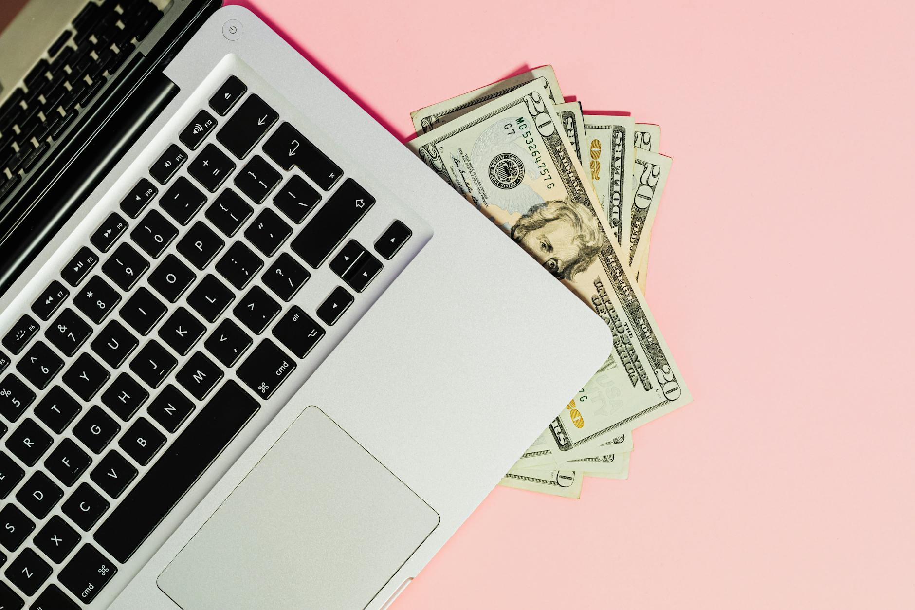 Ilustrasi: A laptop keyboard with dollar bills on a pink surface, symbolizing online business.