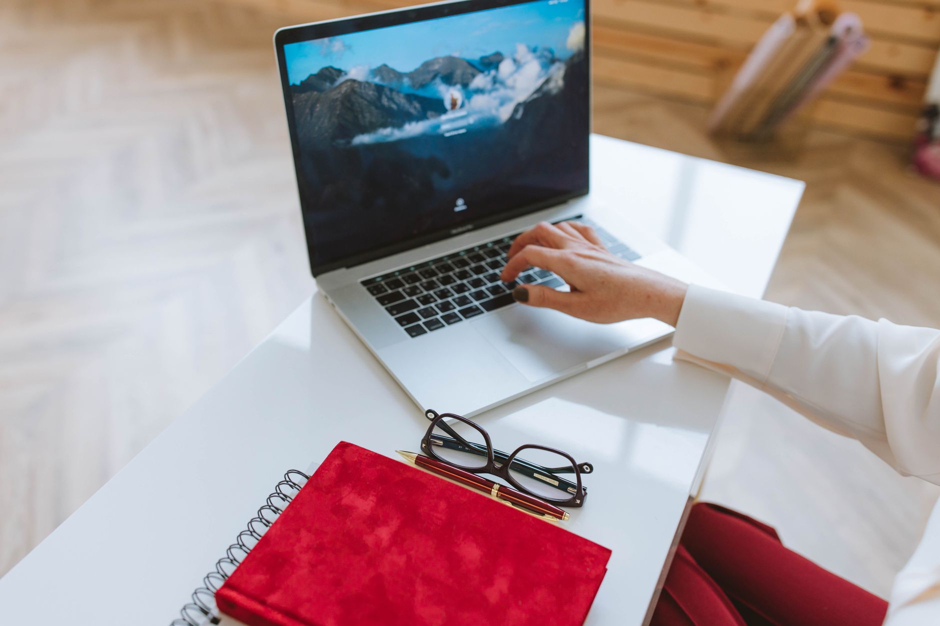 Ilustrasi: A person working on a laptop with a red notebook and glasses on a white table.