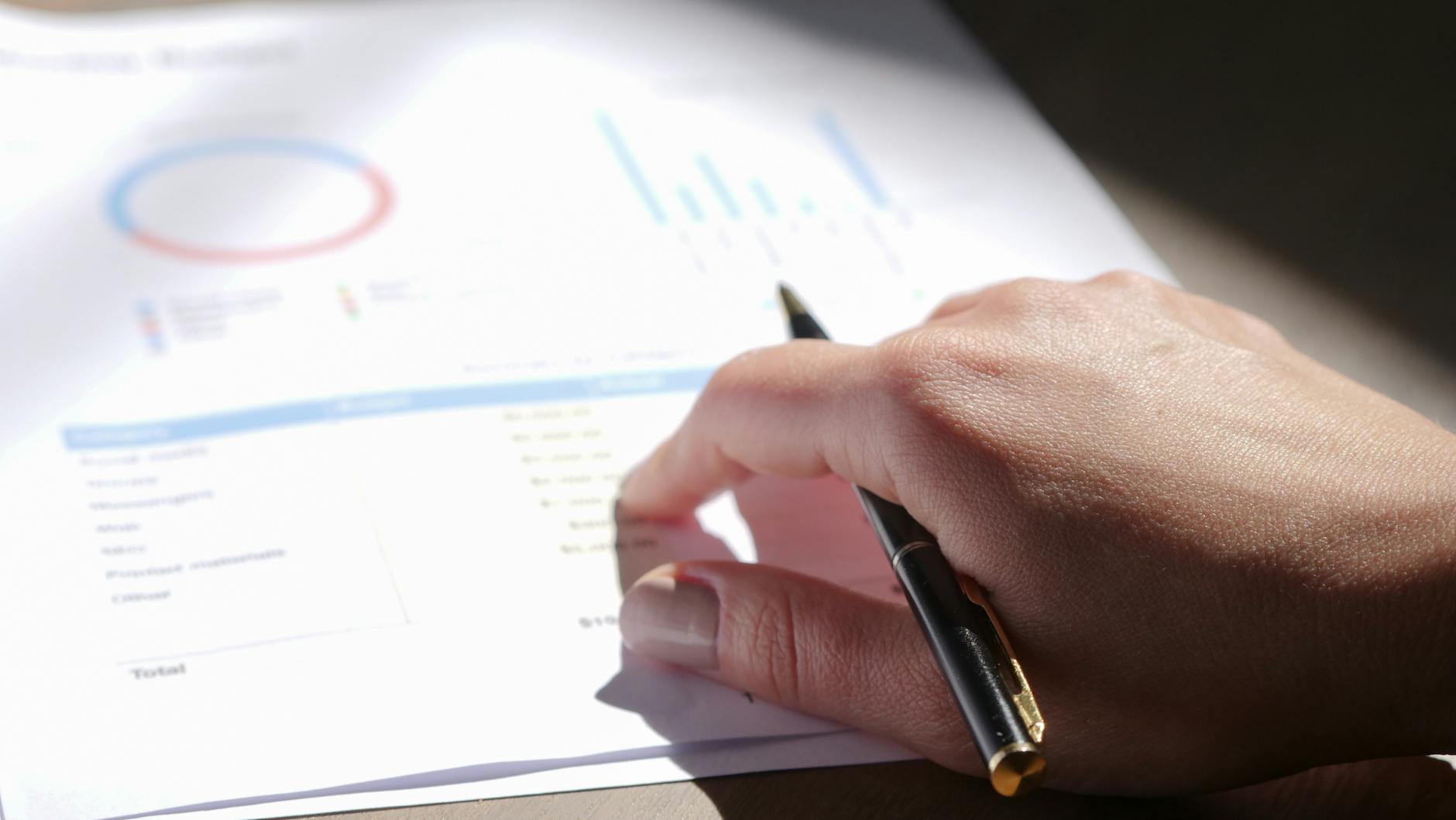 Ilustrasi: A close-up view of a hand holding a pen and examining financial charts on paper.