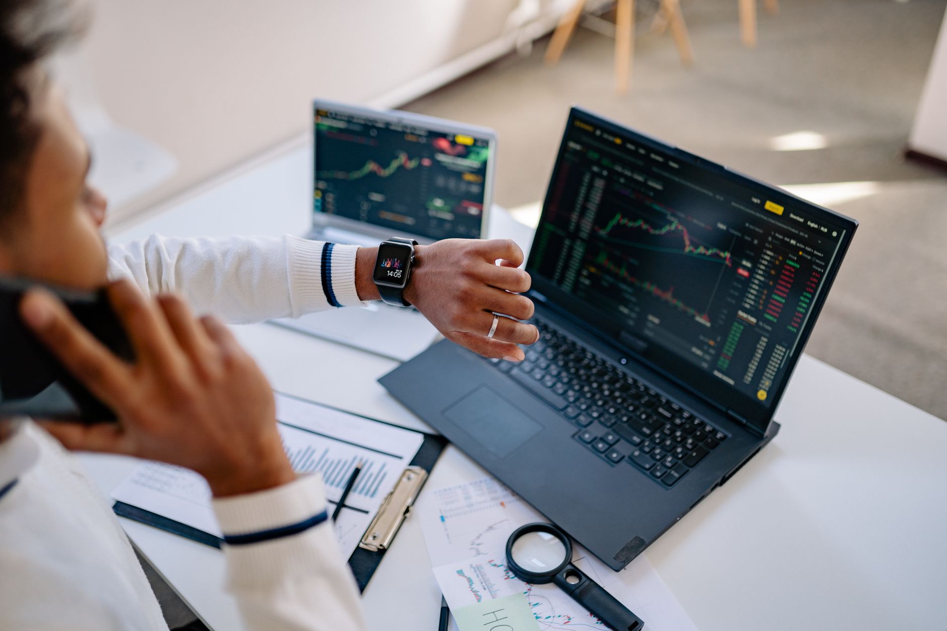 Ilustrasi: Man multitasking with a smartwatch and laptop analyzing stock market charts at the office.