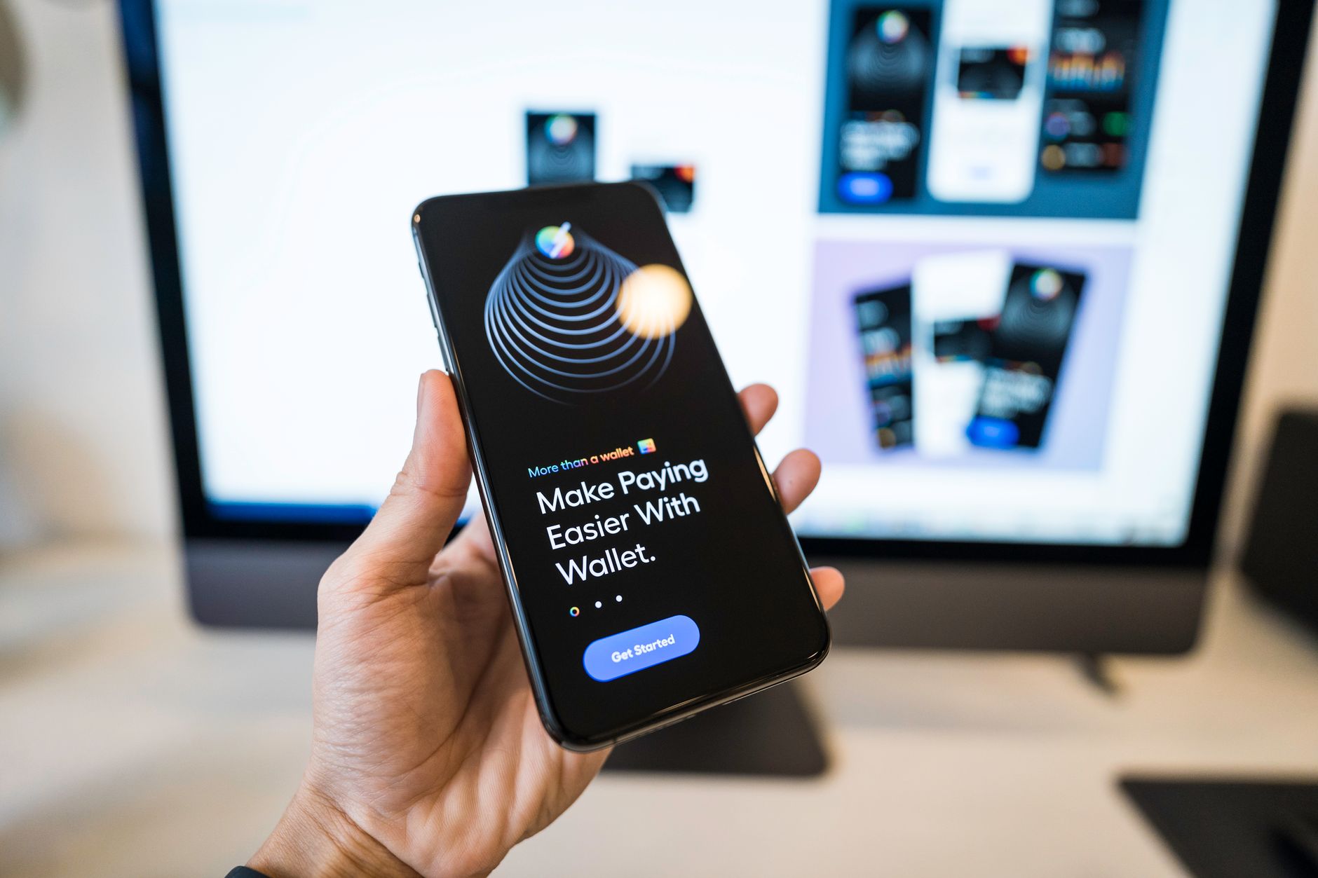 Ilustrasi: Hand holding smartphone displaying digital wallet app interface, blurred monitor in background.