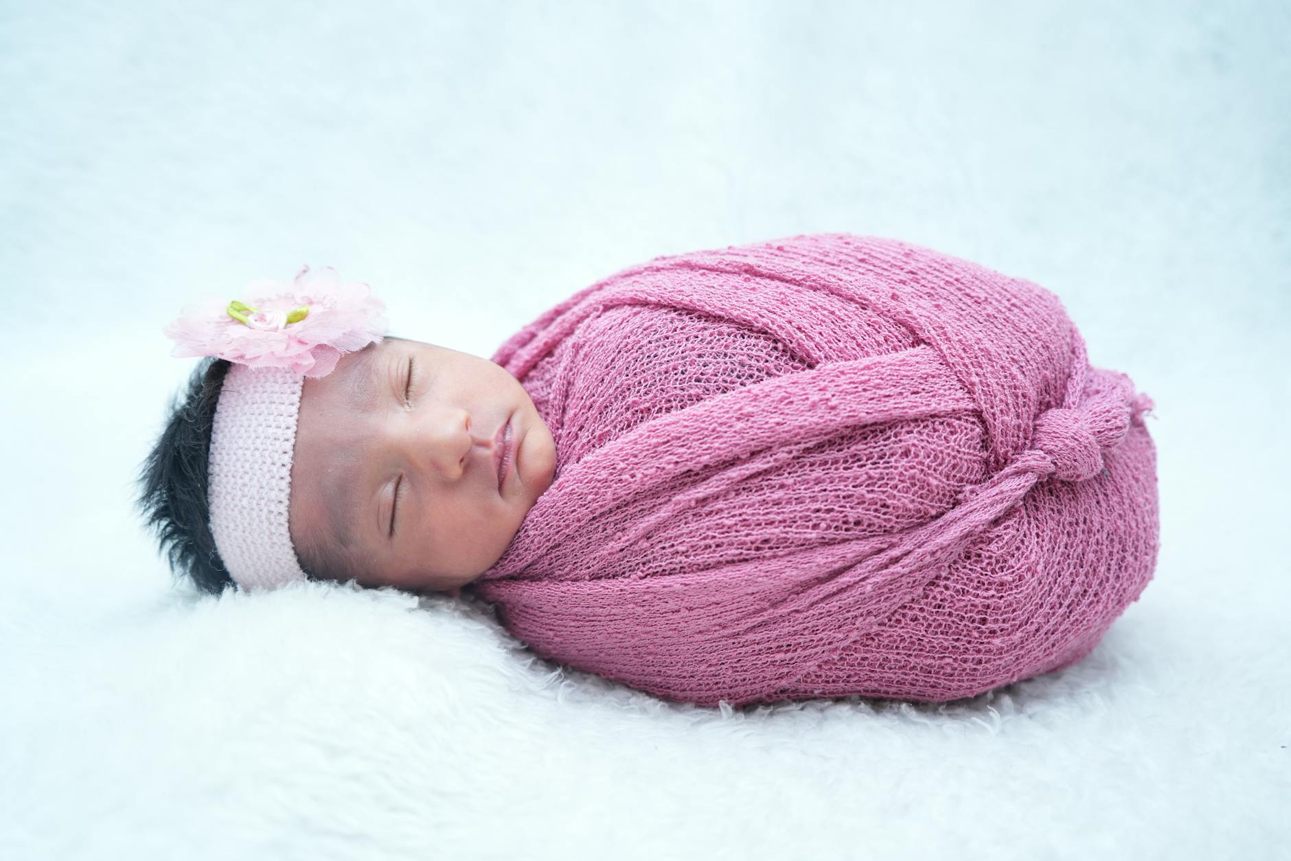 Ilustrasi: A peaceful newborn baby girl wrapped in a pink knitted blanket with a floral headband.