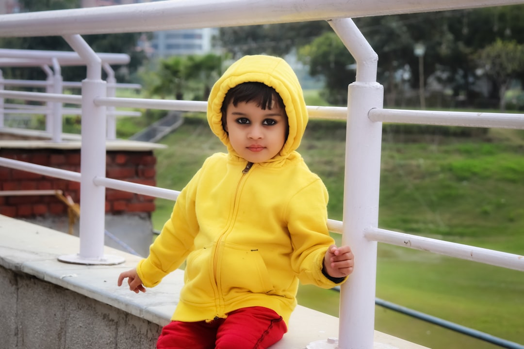Ilustrasi: Child in yellow hoodie jacket and red pants sitting on white metal railings during daytime