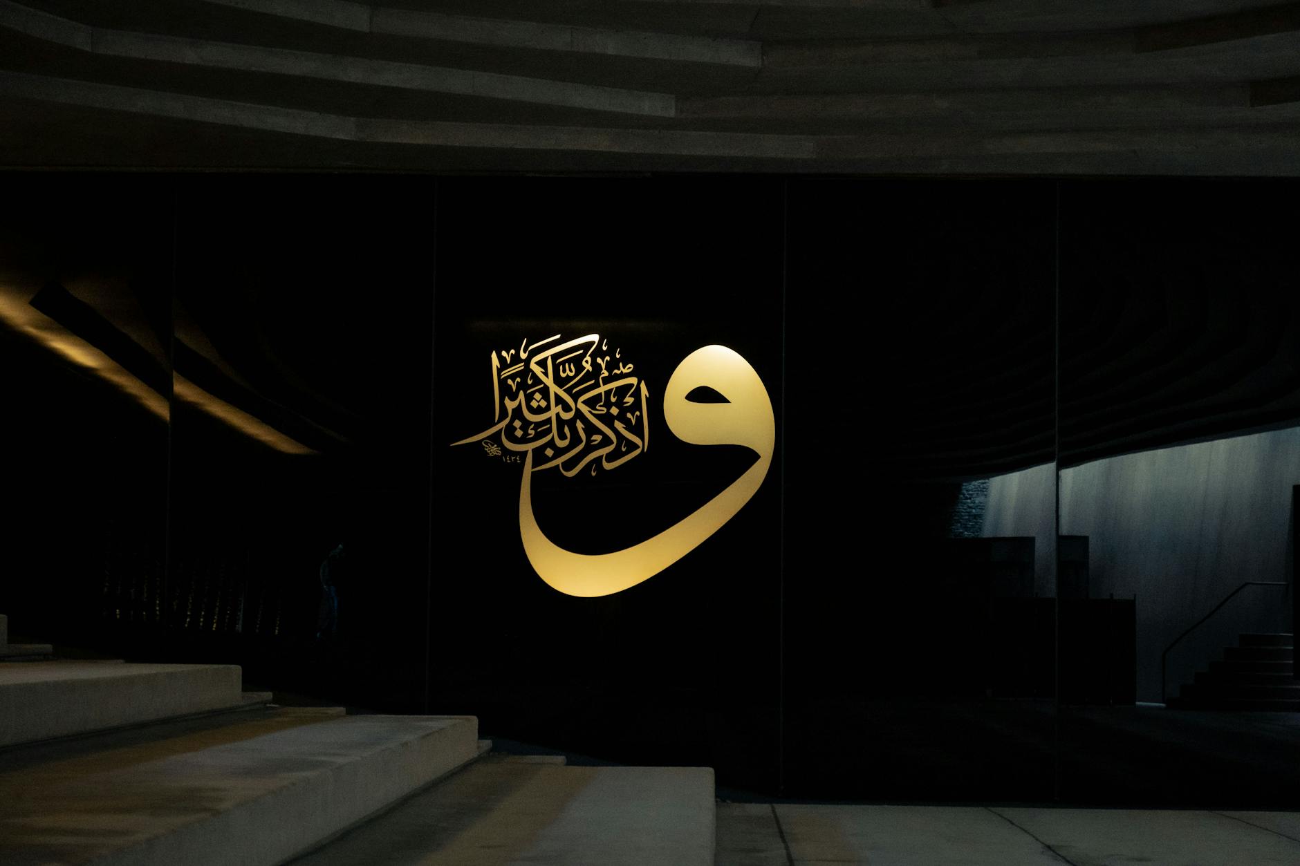 Ilustrasi: Elegant Islamic calligraphy in gold on a modern black wall, featuring architectural elements and stairs.