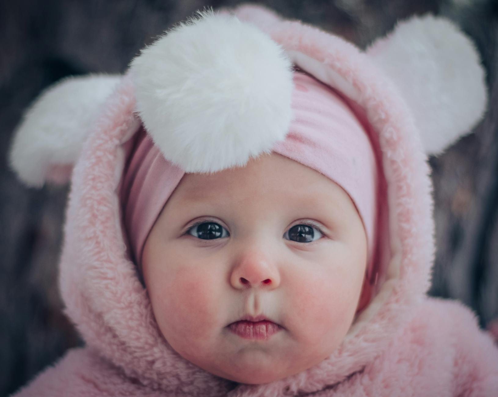 Ilustrasi: Cute baby in a pink fluffy hoodie with adorable pom pom ears. Close-up portrait.