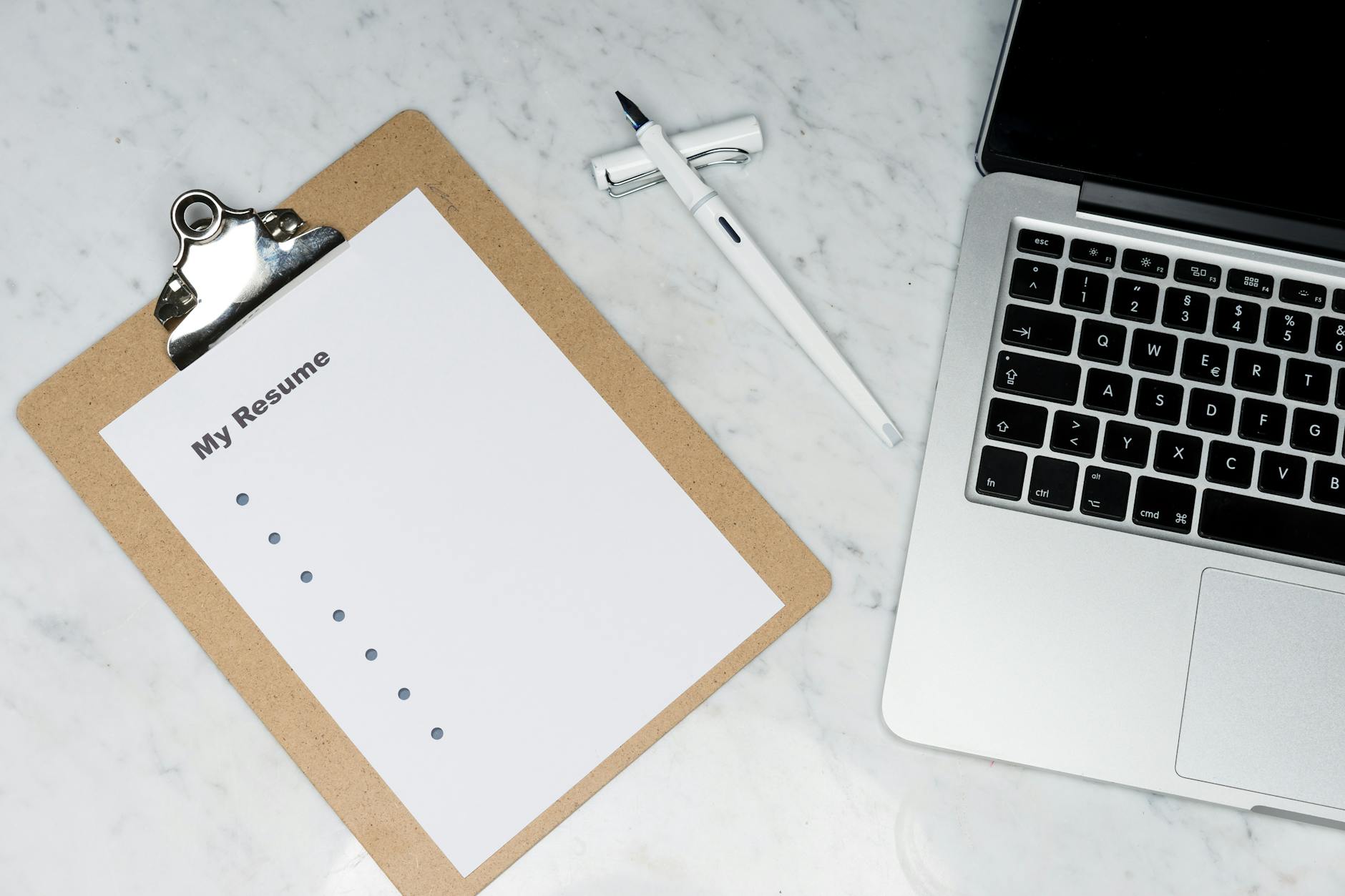 Ilustrasi: A clipboard with a resume beside a pen and laptop on a marble desk, ideal for business and job application themes.