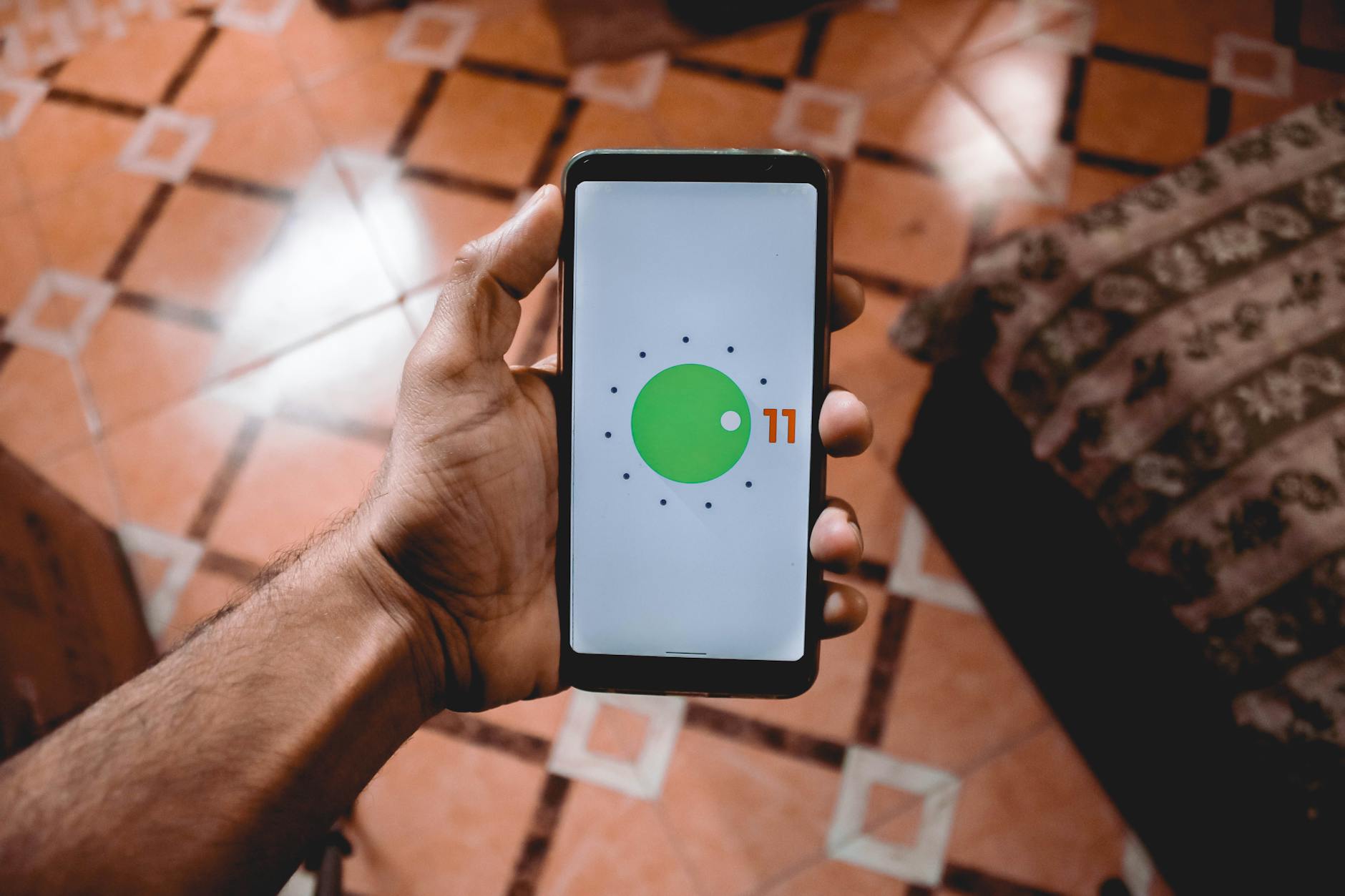 Ilustrasi: Close-up of a hand holding a smartphone displaying Android 11 interface indoors on patterned floor.