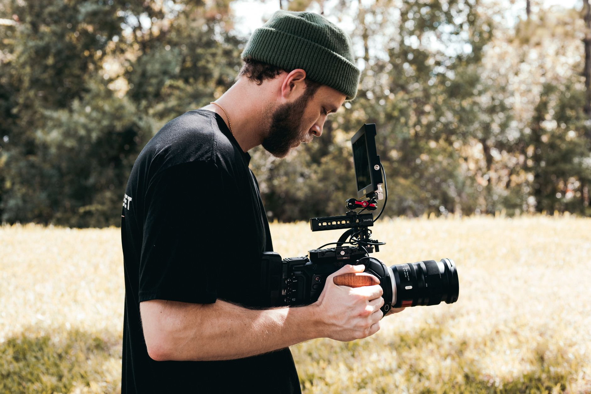 Ilustrasi: Bearded cameraman wearing beanie filming in grassy lapangan with professional camera gear.