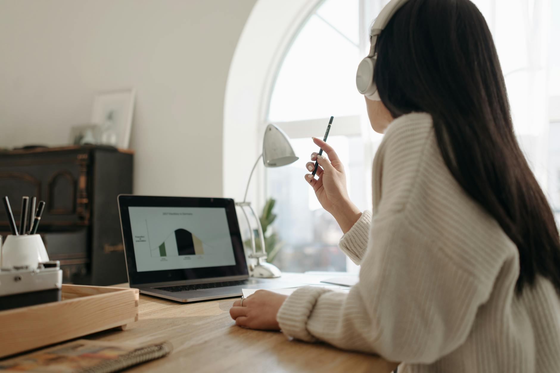 Ilustrasi: Woman with headphones working on laptop analyzing graphs in cozy home office setting. Perfect for remote work visuals.